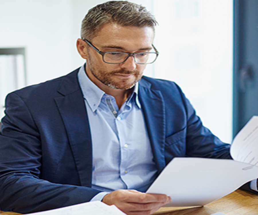 Image of a business man seated at a desk. He is wearing glasses and reviewing a lengthy document, which is secured by a paperclip