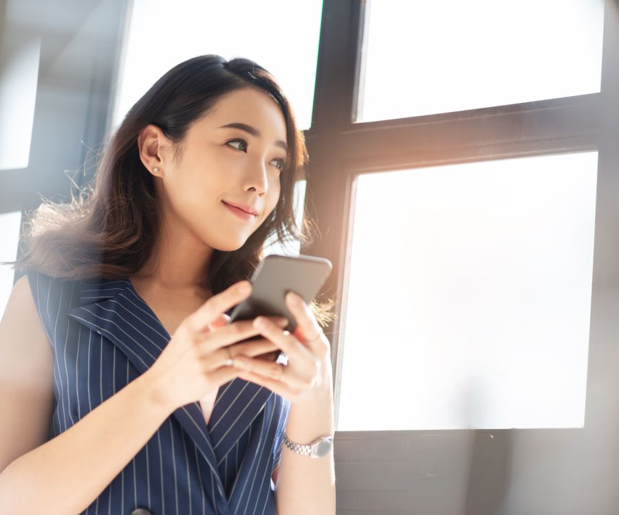 Pleased looking woman using her mobile phone device while standing in front of large light-filled windows