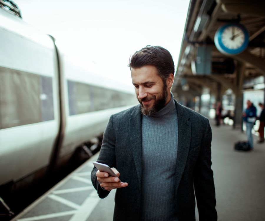 Image of well-dressed middle-aged man standing at station next to a train looking at his phone. It is 2pm.