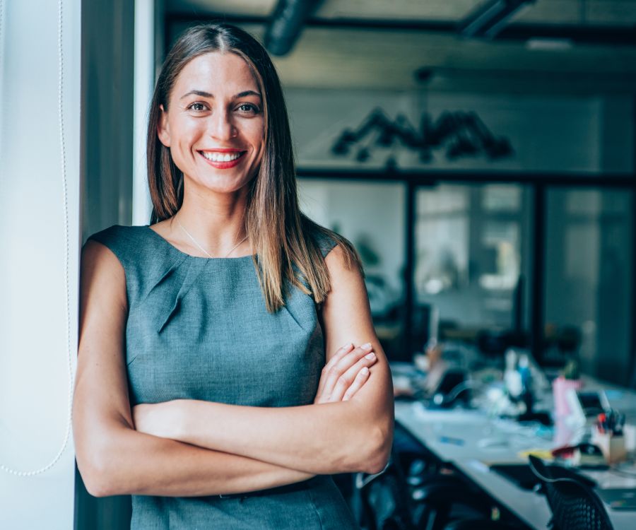 Confident smiling woman leaning against a window frame in a modern meeting room with a table full of papers and laptops