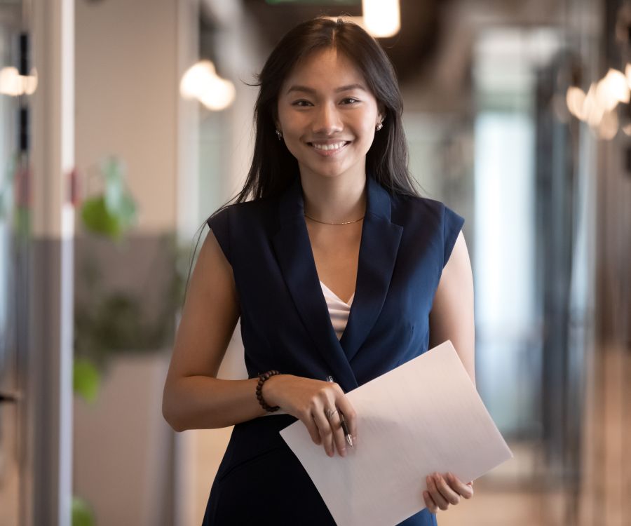 The image shows a smiling professional woman holding papers, standing in a modern office hallway with glass walls and soft lighting.