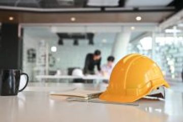 A yellow hard hat rests on a table in a modern office, with people working in the background.