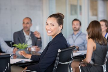 Business people meeting at board table as one woman turns to face the camera and smiles.