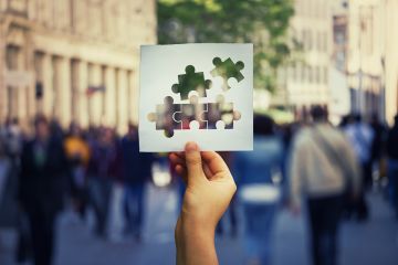 A hand holds a piece of paper with puzzle pieces cut out, with a blurred city street and people in the background.