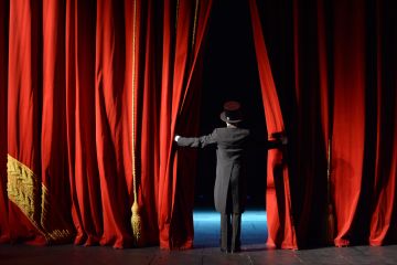 Image depicting rear view of lady in top hat and tails as she peers through parted red curtains to the stage and seats beyond.