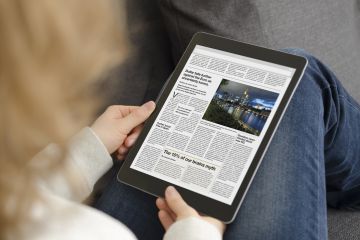 Cropped image of a woman seated sideways on a sofa with tablet resting on her knees. The tablet is showing an online newspaper