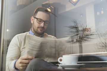 Image of a bespectacled contemporary male enjoying a coffee and reading a newspaper in a coffee shop