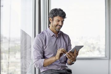 Smiling middle-aged man leaning against office window interacting with a tablet device