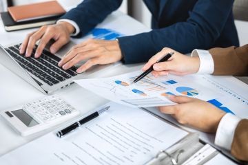 Two professionals are working together, one using a laptop while the other points at a document with charts and graphs, discussing data analysis.