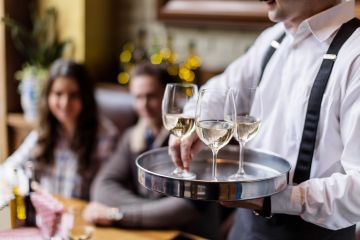 Cropped photo of waiter in white shirt and black braces serving three glasses of white wine to a young contemporary clientele