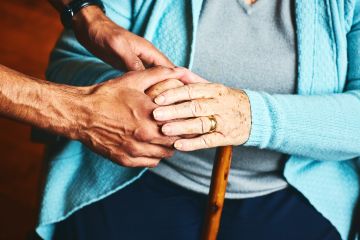 Cropped image of young male hands clasping the folded hands of an elderly seated woman with a walking stick nested on her leg