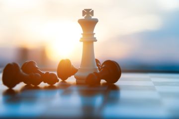 Cropped image of white King chess piece surrounded by five fallen black pawns with a blurred sunset behind.