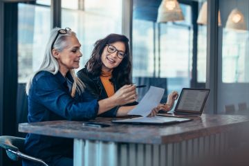 Two female colleagues in an office environment consulting paperwork atop a desk also containing a folder and lap top