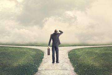 Rear view image of suited man carrying briefcase. He is on a left-right junction of a gravel path. His hand is on his head as though confused