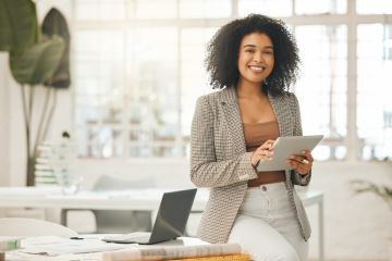 Smiling woman in a blazer holding a tablet, sitting at a desk with a laptop in a bright office.